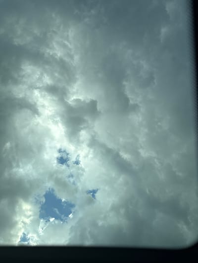 A low-angle shot captures a vast expanse of overcast sky, dominated by a thick layer of grey clouds. Patches of bright blue sky peek through intermittent gaps in the cloud cover, offering glimpses of clear weather. The light filtering through the clouds is diffused, creating a somewhat muted and dramatic atmosphere. The edges of the image are framed by a dark, blurred foreground, suggesting the view is through a window or opening. There are no people, animals, or man-made structures clearly visible in the frame, the focus being entirely on the natural phenomenon of the sky and clouds. The image is devoid of any specific markers that would indicate its location in Dolna Vasilitsa, Bulgaria.