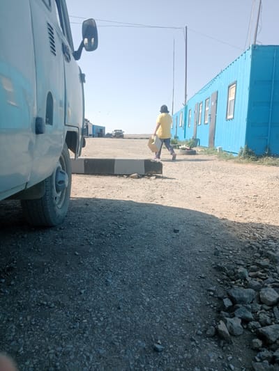 A person wearing a yellow shirt and patterned pants walks on a gravel path in front of a blue metal building with windows and doors. The person is carrying a cardboard box. To the left of the path, a light-colored vehicle is partially visible. The scene is outdoors with a clear sky. The location is in Қарасу ауылдық округі, Kazakhstan.