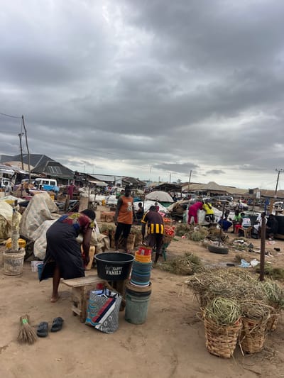 The image depicts a bustling outdoor market scene in Oko Amakom, Nigeria, under an overcast sky. In the foreground, a woman wearing a colorful patterned dress and a dark skirt is bent over, tending to goods on the ground. Next to her, a large black plastic basin with the inscription "HAPPY FAMILY" sits on a wooden stool. A variety of stacked colorful plastic containers and buckets are visible. A broom and a pair of black sandals rest on the sandy ground to the left.
Further into the market, several individuals are engaged in commercial activities. One man in an orange tank top with a graphic print stands facing the camera, while another man in a yellow and black striped shirt is partially visible. Baskets filled with produce, likely tomatoes, are prominently displayed in woven containers. Various tents, makeshift stalls, and vehicles, including a blue van, form the market infrastructure. Bundles of dry grass or fodder are spread out on the ground, and some are also contained within baskets. The overall environment suggests a lively and active marketplace.