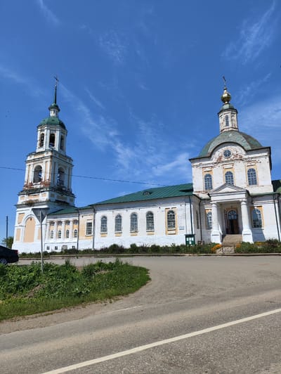 The image shows the Church of the Resurrection in the village of Mostovoye, Russia. The church is a white brick building with a green roof. It has a tall bell tower on the left and a dome on the right. The sky is blue with some white clouds. There is a road in the foreground with a car parked on the left side of the image. The church appears to be in good condition, with some signs of age on the bell tower. The overall scene is peaceful and serene.