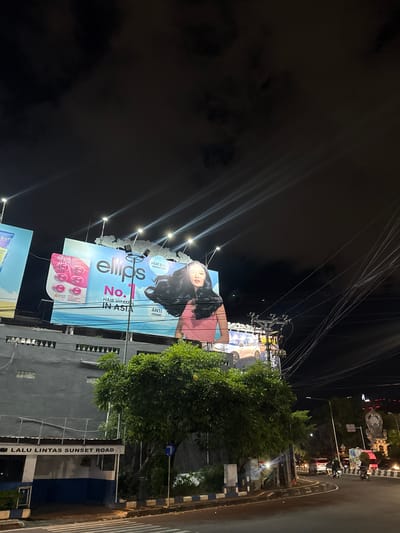 This media file captures a night scene in Denpasar Barat, Indonesia, featuring a large billboard advertising "ellips Hair Vitamin." The billboard displays a woman with long dark hair and the product packaging. The text on the billboard highlights "Heat & UV Protection" and "Anti Oxidant," along with the claim "No. 1 Hair Vitamin in Asia."
The setting appears to be a street, with overhead power lines, streetlights, and a road visible. The time of day is clearly night, as indicated by the dark sky and the illuminated billboard. The presence of a street sign reading "LALU LINTAS SUNSET ROAD" provides a specific location cue. A lush green tree is situated in front of a building behind the billboard, partially obscuring it. The overall mood is commercial and urban, with the bright billboard contrasting against the night sky.