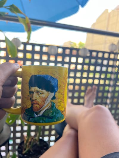 A person's hand holds a yellow mug adorned with a depiction of Vincent van Gogh's self-portrait. The mug is positioned in front of a blurry outdoor scene, featuring a black metal table with a grid pattern and the legs of a person sitting at it. Sunlight streams in from the left, illuminating part of the scene and creating lens flare. In the background, out-of-focus greenery and shadows suggest a garden or patio setting. The image appears to be taken during the daytime, with bright sunlight indicating it's likely morning or afternoon. The overall mood is relaxed and casual.