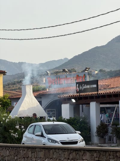 In Santu Diadòru/San Teodoro, Italy, a white Peugeot car is parked in front of a building with a red-tiled roof. Smoke is rising from a white, conical chimney. A sign above the entrance reads "Dettorimarket" in red lettering, and another black sign displays "La Gallurese Gastronomia" with an illustration of a chef. Behind the building, a wooded hillside and mountains are visible. A person is partially obscured by greenery near the chimney. Clothing is displayed for sale on racks outside the Gastronomia. The scene is framed by two parallel power lines in the foreground and a stone wall.