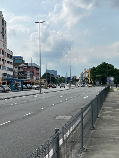The image depicts a street scene in Subang Jaya City Council, Malaysia, under a cloudy sky. A multi-lane road dominates the foreground, with white lane markings. A metal fence runs along the right side of the road, bordering a concrete sidewalk.
On the left side of the road, a row of buildings is visible. The closest building is a multi-story residential complex with balconies and windows. Adjacent to it is a row of shops with blue awnings and parked cars. Further down the road, more buildings of varying heights and architectural styles are present, including a building with a distinctive red and white facade and what appears to be laundry hanging from balconies.
Traffic on the road includes several cars, a yellow vehicle, and what appears to be a motorcycle. Some vehicles are parked along the side of the road near the bus stop. Traffic lights are visible at an intersection in the distance.
Streetlights are a prominent feature, with tall poles equipped with dual lights positioned along the road. Green trees are present on the right side of the road, behind the buildings, and in the distance, with more modern high-rise buildings visible further away. A few people are observed on the sidewalk and near the bus stop. The overall impression is of a busy urban environment on a slightly overcast day.