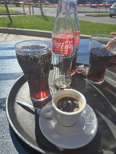 The image depicts a sunlit outdoor scene in Cars, France. Centered on a dark, textured serving tray are two tall glasses containing a dark, carbonated beverage. Between these glasses stand two clear glass bottles of Coca-Cola, each displaying a red "Coca-Cola GOÛT ORIGINAL" label. In the foreground, a white ceramic espresso cup filled with dark coffee and crema is placed on a matching saucer, alongside a small metallic spoon. An opened wrapper, consistent with sugar or candy, is visible near the right glass. A partial human hand is visible at the right edge of the frame, positioned near the rightmost glass. The background reveals a paved area, a bright green grassy verge, and a road with a partially visible white car. Distant bare trees or bushes are also discernible under clear, sunny conditions.