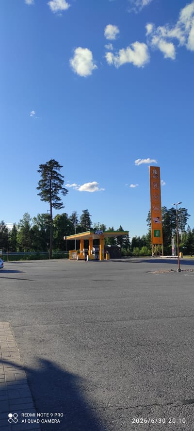 This media file shows an outdoor scene at a gas station in Joutsa, Finland. The image is oriented vertically, with the sky occupying the left side and the gas station and road on the right.
The sky is a vibrant blue with scattered white, fluffy clouds. The sun appears to be shining brightly, suggesting it is daytime. The gas station itself is brightly colored in yellow and orange, with a prominent "ABC!" sign. Gas pumps are visible under the covered area. Trees, mostly coniferous, form a backdrop behind the gas station. A tall, slender pine tree with dense foliage stands in the foreground on the left side, reaching towards the sky.
The road in front of the gas station is paved with asphalt. A small shadow from an unseen object falls across the road on the lower right. In the upper right corner, a timestamp "2026/6/30 20:10" and text related to a "REDMI NOTE 9 PRO AI QUAD CAMERA" is visible, indicating the image was captured with a mobile phone. There are no people clearly visible in the image. The overall impression is a sunny, clear day at a functional, modern gas station in a somewhat rural or wooded setting.