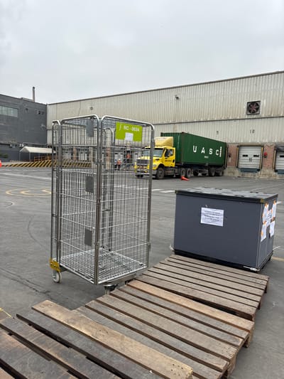 This is a photograph taken outdoors at what appears to be a shipping or logistics yard. The scene is dominated by industrial structures, including a large building with corrugated metal siding and loading bay doors. A green shipping container, marked with "UASC," is being transported by a yellow truck, also labeled "RANSA." Next to the truck, a large dark gray container rests on a wooden pallet. In the foreground, a metallic cage trolley with wheels is positioned.

Visible text includes numbers on the building (9, 10), the shipping container's branding "UASC," and the truck's branding "RANSA." There is also a label on the dark gray container with Spanish text that appears to relate to shipping information or incident reports: "PEDIDOS NO SHOW CON INCIDENCIA FLB CAYMA-C&C F. ENVIO: 25.06.26." Another label on the cage trolley reads "RC-0634."

The time of day appears to be daytime, with overcast or cloudy weather indicated by the diffused light. The location is suggested to be Villa El Salvador, Peru, based on the provided context, although this is not directly visible in the image itself. The overall atmosphere is industrial and functional, with no people clearly visible in the frame. The focus is on the cargo, vehicles, and equipment associated with transportation and warehousing.