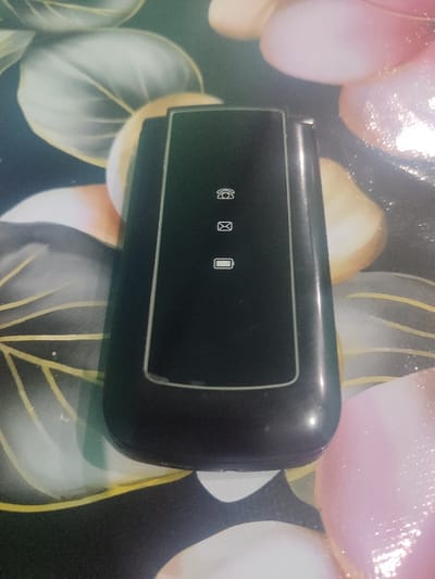 A closed black flip phone rests on a patterned surface with flower-like designs. The phone's exterior features icons for phone call, email, and battery level. The surface appears to have a glossy finish, creating reflections. The background consists of stylized floral patterns in shades of white, pink, yellow, and gray.