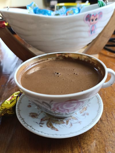 A close-up, eye-level shot showcases a traditional cup of coffee on a saucer, placed on a wooden table. The coffee is a rich brown with a frothy top, suggesting it's freshly brewed. The cup and saucer are adorned with a delicate floral pattern. To the left of the cup, a golden candy wrapper is visible. In the blurred background, a white ceramic bowl is filled with various colorful candies. The overall scene is warm and inviting, hinting at a moment of indulgence and relaxation in Adzina, Abkhazia.