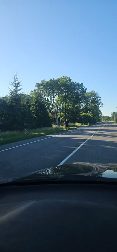 The image shows a person running along the side of a road in Tuulna, Estonia. The road is asphalt with white lane markings. The person, wearing a dark t-shirt and shorts, is running on the right side of the road, facing away from the camera. To the left of the road, there is a grassy shoulder with tall grass and scattered trees, including a tall spruce tree and several large deciduous trees. In the background, there are more trees and a clear blue sky. The foreground of the image shows the dashboard of a vehicle, with the reflection of the sky and trees visible on its surface. There are also road signs visible on the side of the road. The lighting suggests it is daytime, likely late afternoon or early morning due to the long shadows cast by the trees.