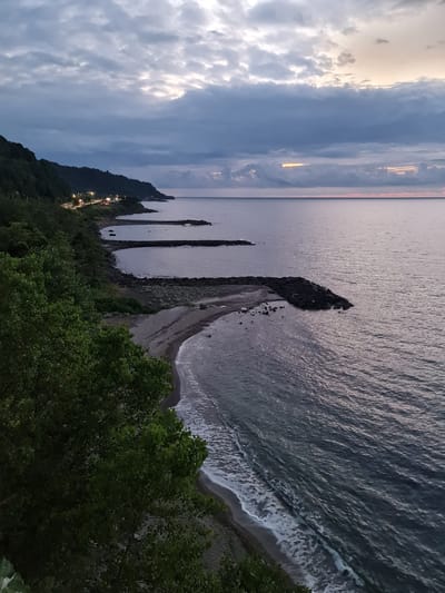 The image depicts a coastal scene at dusk in Beşikdüzü, Turkey. The sky is overcast with dramatic clouds, illuminated by the faint glow of twilight and distant lights. A dark, tree-covered hillside lines the left side of the frame, with a road visible running along its base, dotted with streetlights and vehicle lights. Several rocky breakwaters extend from the shore into the calm, grey sea, creating small, protected coves. The beach is composed of pebbles and sand, with gentle waves lapping at the shore. The overall atmosphere is serene and slightly melancholic, characteristic of a coastal evening.