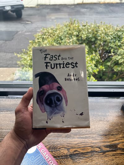 A person holds a book titled "The Fast and the Furriest" by Andy Behrens. The book features a close-up image of a dog's face on the cover. In the background, there is a window with a view of greenery and an overcast sky. A second book is partially visible to the right, with the words "CK GANTO" visible on the cover. The photograph was taken in the Town of New Windsor, United States of America.