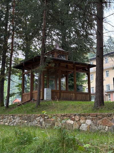 A wooden gazebo with glass windows is situated on a grassy slope behind a stone wall. A light-colored electrical box is mounted on the exterior of the gazebo. In the background, a multi-story building with yellow siding and white windows stands. The scene is surrounded by tall trees, with pine trees being prominent. The location appears to be in Świeradów-Zdrój, Poland, possibly in a park or recreational area.