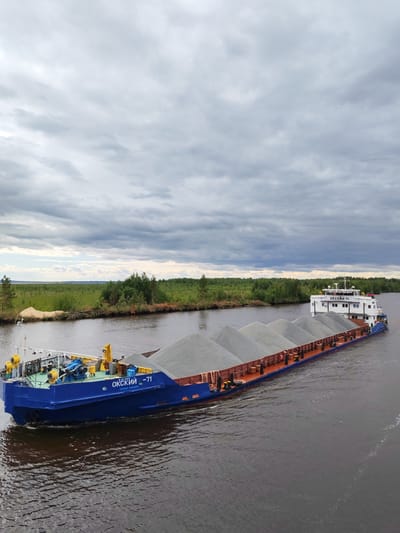 A blue barge, identified by the Cyrillic text "ОКСКИЙ -71" on its side, is navigating a dark, calm river under a cloudy sky. The barge is laden with a substantial cargo of light gray gravel, piled high in multiple conical mounds. The barge has a white superstructure with multiple windows and antennae at the stern. The riverbanks are lined with dense green trees and vegetation, with a patch of sandy soil visible on the left bank. The overall scene suggests a calm day on a waterway, likely for transporting bulk materials. The location is specified as Ozernoe Ust'e, Russia.