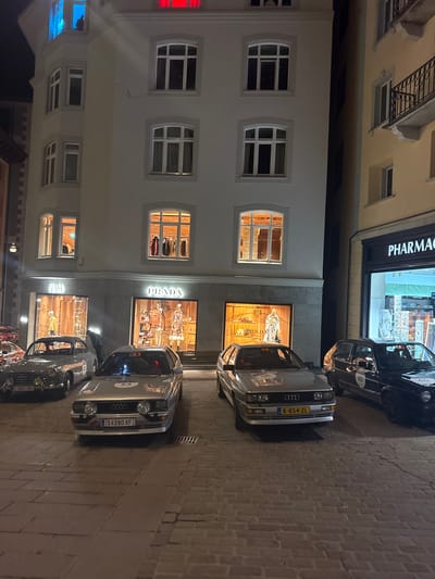 This nighttime image depicts a street in Sankt Moritz, Switzerland, showcasing vintage rally vehicles parked on a wet cobblestone surface in front of illuminated buildings.

The central structure is a multi-story, light-colored building. Its ground floor houses a Prada boutique, identifiable by its prominent signage and multiple display windows exhibiting mannequins, clothing, and accessories. Upper floors feature arched windows; one on the first floor displays clothing racks, while a corner turret window is illuminated with a blue light, revealing a silhouetted figure. A top-floor window shows a red internal light. Adjacent to the right, a yellow-toned building features a 'PHARMACY' on its ground level and an upper-floor balcony with an iron railing. A dark, stone-faced building is partially visible on the far left.

Four distinct classic/vintage cars, each marked with a white circular rally number, are parked parallel to the Prada building. From left to right:
1.  A dark grey/silver vintage coupe or convertible (German plate "D-AHX-081", number "52") equipped with yellow fog lights.
2.  A silver Audi Quattro (Swiss plate "S-280 XF", number "53").
3.  A silver Audi Quattro (Dutch plate "K-854-ZL", number "58").
4.  A black Volkswagen Golf hatchback (number "60").

One individual is partially visible, bent over between the vintage coupe and the Audi "53". The overall illumination originates from interior building lights and storefronts, casting reflections on the damp cobblestone street.