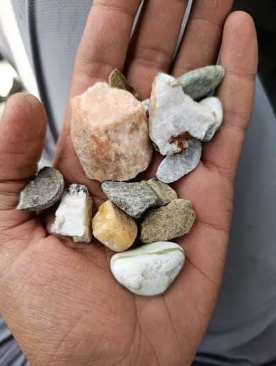 A person's hand, displaying visible lines and texture, holds a collection of small rocks and stones. The rocks vary in color and shape, including shades of white, pink, grey, yellow, and green. Some are rough and crystalline, while others are smooth and rounded. The background appears to be a grey fabric. The context of Khuroson, Tajikistan, is not discernible from the visual information provided in the image.