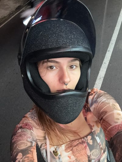 A close-up, top-down shot features a person wearing a black full-face helmet. The helmet has a matte finish with what appears to be glitter speckles on the exterior. The person's face is visible through the helmet's opening, displaying light brown eyes and a silver nose ring. They wear a long-sleeved shirt with a floral pattern in shades of pink and brown. The background consists of asphalt, with a white painted line partially visible in the upper-left corner. The location is identified as Tumbak Bayuh, Indonesia.