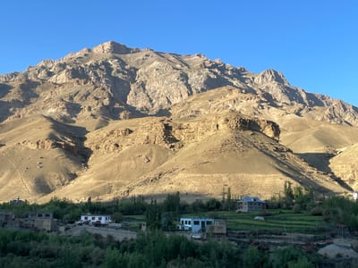 The image depicts a remote village nestled at the base of a large, barren mountain range under a clear blue sky. The mountain's rugged, rocky terrain is illuminated by sunlight, casting shadows across its slopes and revealing layers of geological strata. In the foreground, the village consists of several houses with traditional architecture, characterized by flat roofs and stone or mud-brick construction. Some houses are clustered together, while others are more isolated. Lush green vegetation, including trees and cultivated fields, surrounds the village, creating a stark contrast with the arid mountains. A road runs through the village, with a few vehicles visible, including a white truck and a car. One of the houses is painted white with a blue trim. In the distance, a domed structure, possibly a stupa or a mosque, is visible on a hillside. The overall scene conveys a sense of isolation and peaceful rural life within a dramatic natural landscape. The location is identified as Khangkor, India.