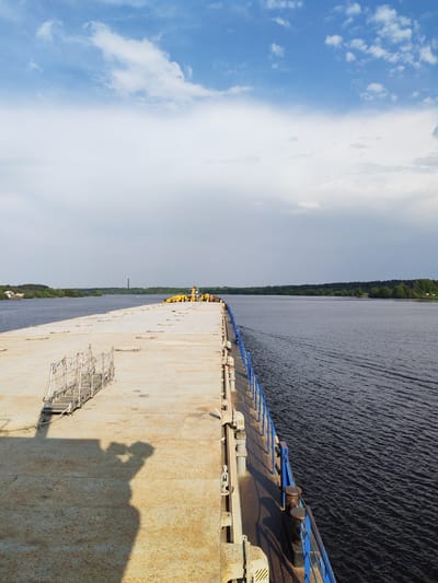 The image depicts a long, flat barge on a body of water, likely a river or lake, under a partly cloudy sky. The barge extends from the lower left corner towards the center, with its deck appearing to be made of metal and showing signs of wear. A short set of stairs with a metal frame is visible on the left edge of the barge. A blue railing runs along the right side of the barge, separating the deck from the water. In the distance, a line of trees is visible on the shore, with a few structures and a chimney-like structure peeking above the trees. The water surface is slightly rippled, indicating a gentle breeze. The shadow of the photographer is visible in the lower-left corner of the image. The location is identified as Nutromo, Russia.