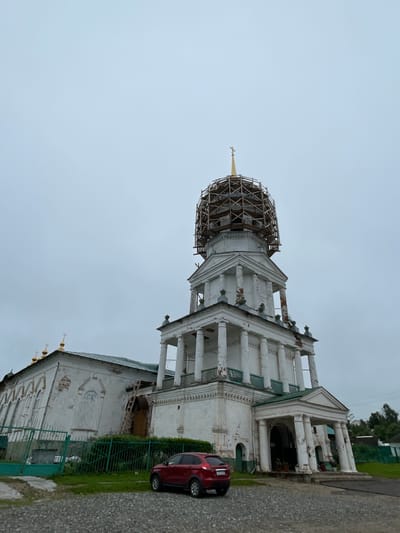The image shows a white, multi-story church with a green roof and a golden cross on top. The church appears to be under renovation, as scaffolding is visible around the upper dome. A red car is parked in front of the church. The sky is overcast. The church is located in the village of Кстинино, Russia.