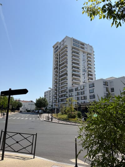 This is a vertical, eye-level, medium shot of a street in Courbevoie, France, on a sunny day. The scene depicts a modern urban environment with large residential buildings and a street.

The dominant subjects are the tall, white, multi-story apartment buildings with numerous balconies and windows. One building is particularly prominent in the foreground, featuring a stepped architectural design. On the right side of the image, a street with visible lane markings and a pedestrian crossing leads away into the distance. A black metal fence lines the sidewalk. Green trees and foliage frame the upper portion of the image and are also visible along the street.

In the distance, other buildings of varying heights can be seen. There are also streetlights, traffic signs, and a few parked cars. A black directional sign is mounted on a pole in the foreground.

The weather appears to be clear and sunny, indicated by the bright blue sky and the strong shadows cast by the trees and fence. The time of day is likely daytime, given the bright sunlight. There are no discernible people in the frame.

Visible text includes "HAPPY HOURS 7PM THRU 9PM" and "PASTRERIE" on a sign attached to a building on the left side of the street. There are also some smaller, illegible signs on poles and buildings. The overall impression is one of a well-maintained, modern residential area in a French city.