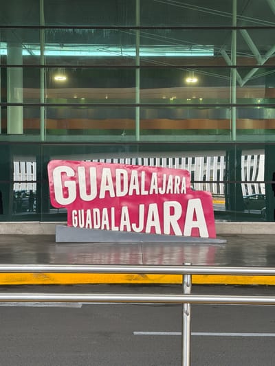 A sign displays "GUADALAJARA" in bold white letters on a red background, repeated in smaller font below. The sign sits beside a modern glass building, reflecting light. A yellow-painted curb and metal guardrail edge a dark asphalt surface marked with white lines. The shot has been rotated.