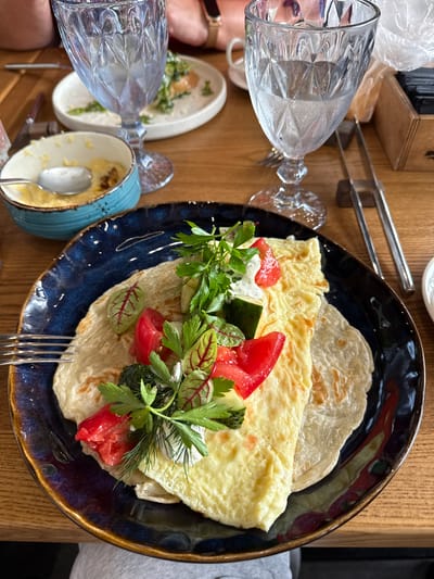 A close-up, overhead shot captures a table setting at a restaurant in Nizhnekayancha, Russia. The main focus is a dark blue, artisanal plate holding a breakfast dish. The dish consists of thin, layered crepes topped with a folded omelet, fresh chopped tomatoes, cucumber slices, and a sprinkle of mixed greens. A dollop of white sauce or cream is visible, along with fresh dill and parsley garnishes. To the left, a light blue ceramic bowl with a spoon contains a yellow, semi-liquid food, possibly a sauce or a dip. In the background, two tall, faceted glasses of water are placed on a wooden table. One glass is mostly full, with condensation visible, while the other is nearly empty. The table also features chopsticks in a wooden holder on the right side. The overall impression is of a leisurely meal at a casual dining establishment.