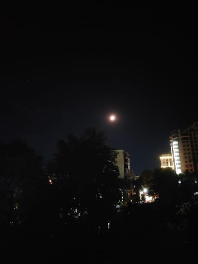 A nighttime, low-light photograph captures a section of the city of Tursunzoda, Tajikistan, under a dark, cloud-streaked sky. The dominant celestial body is a bright, full moon, appearing as a luminous disc positioned in the upper left quadrant of the frame. Below the moon, silhouetted trees and vegetation form a dense, dark foreground.

To the right of the trees, two multi-story buildings are visible. The building closer to the center of the frame is illuminated, showing rows of windows, and appears to be a residential or office complex. Further to the right, a taller, unfinished building stands, its facade also lit, revealing construction elements and multiple windows. In the distance, between the buildings and trees, is a cluster of lights suggesting a more populated area or street scene, possibly indicating a lively part of the city. The overall impression is one of urban life continuing under the night sky, with the moon casting a soft glow.