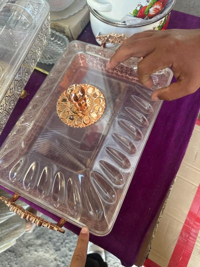 An overhead shot shows a person's hands interacting with a clear plastic serving tray with rose gold handles and a rose gold decorative element in the center. The tray has individual compartments shaped like teardrops. To the left of the serving tray is another clear plastic container with ornate rose gold trim. The items are placed on a dark purple velvet cloth, which is on top of a light-colored, textured surface. In the background, a white and red patterned pot with strawberries is visible. The scene appears to be set in an outdoor market or stall, possibly in the city of Kara-Suu, Kyrgyzstan, known for its bazaars. The lighting is natural, suggesting daytime.