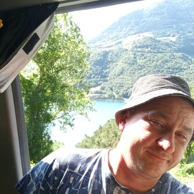 A man wearing a bucket hat takes a selfie from inside a vehicle, looking towards the camera with a slight smile. The background reveals a scenic view of a body of water, likely a reservoir, nestled within lush green mountains. The sky is bright and appears to be daytime. The interior of the vehicle is visible on the left side of the image, with a black sunshade and a grey seatbelt strap. The man is wearing a blue and white patterned t-shirt. The location is identified as Corps, France, suggesting a mountainous and possibly rural setting within that region.