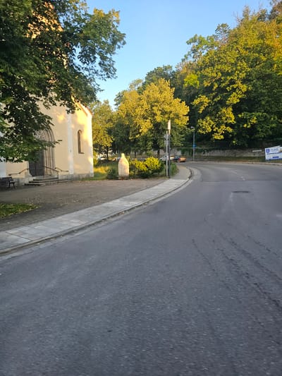 The image depicts a street scene in Pappenheim, Germany, likely during the late afternoon or early evening due to the warm sunlight illuminating the trees and building. On the left side of the frame is a light-yellow building, possibly a church or public institution, with a dark arched doorway and a single arched window. In front of the building, there is a paved walkway with a dark bench. Adjacent to the walkway is a statue of a robed figure. The street itself is paved and curves to the right, with a sidewalk separating it from the walkway. Lush green trees line the upper edge of the scene, casting shadows on the road and buildings. In the distance, a stone wall runs along a wooded hillside. A few cars are visible on the road, including one white car and a darker sedan. A blue parking sign is also present. A large white billboard with German text, including "170 JAHRE", is situated in the background on the right side of the road. The sky is clear and blue.