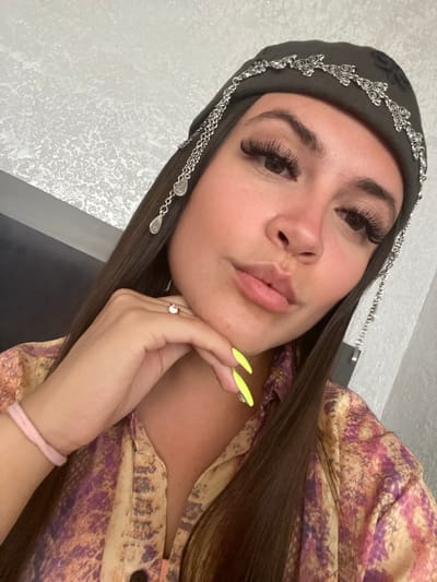 A young woman with long, dark hair is shown from the chest up, looking towards the camera with her chin resting on her hand. She is wearing a dark beanie adorned with silver chain detailing and teardrop-shaped charms. Her fingernails are painted a bright neon yellow. She is also wearing a patterned shirt with shades of purple, pink, and yellow. A thin pink bracelet is visible on her wrist, and she has a ring on her right ring finger. The background appears to be an indoor setting with a textured white wall. There is no information in the image to suggest it is located in Chalda, Russia.