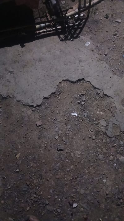 The image depicts a ground surface with a distinct border between concrete and loose dirt/gravel. The concrete section is on the left side of the image and appears weathered with cracks and an uneven surface. The right side is comprised of a darker, granular material, possibly a mix of dirt, gravel, and small stones. Scattered debris is visible on both surfaces, including what appears to be a small piece of white paper or plastic. Part of a metal structure (possibly a cage or frame) is visible in the lower-left corner, casting a shadow. No people are visible. Given the reference to Putucual, Venezuela, the scene is likely an outdoor area in that location, possibly a yard or pathway.