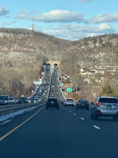 The image captures a multi-lane highway experiencing traffic congestion during daylight hours on a winter day. The scene is viewed from inside a vehicle, looking ahead down the road.
In the immediate foreground, a silver SUV is prominent in the right lane, with its rear displaying a license plate that reads "111-WYL". A black sedan follows directly behind it, and other vehicles are visible further back in the same lane. To the left, an adjacent lane is also occupied by numerous cars in traffic, separated by a concrete barrier.
The highway curves gently into the distance, leading to two distinct tunnels. These tunnels are characteristic of the Heroes Tunnel (formerly West Rock Tunnel) on CT Route 15. Above the highway near these tunnels, green road signs are visible, one of which clearly displays "EXIT 59".
To the left of the highway, a steep hillside covered with bare deciduous trees and scattered patches of snow rises, indicating a winter or early spring environment. A tall cellular or radio tower stands on this hillside. On the right side of the image, beyond the highway and wooded areas, several residential or commercial buildings can be seen.
The sky is a bright blue, adorned with several elongated white clouds. The overall impression is one of cold weather and heavy vehicular traffic, likely causing delays. Given the context of New Haven, United States, and the visible "EXIT 59" and twin tunnels, the location is identifiable as the Heroes Tunnel area near New Haven, Connecticut.