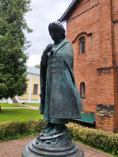 The image shows a bronze statue of a person in historical attire, standing on a pedestal in front of a red brick building. The statue depicts a figure wearing a patterned robe, a hat, and gloves. The building behind the statue has arched windows and decorative brickwork along the roofline. To the left of the statue, there is a tall evergreen tree and a yellow building with white trim. The ground is paved with red bricks, and there is a well-maintained lawn and bushes surrounding the statue. The sky is overcast. This scene is located in Chelganovo, Russia.