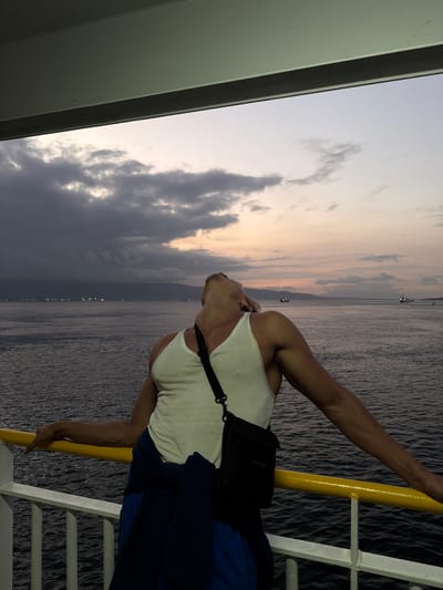 A man with tanned skin, wearing a white tank top and blue shorts, leans back with his head tilted upwards, gazing at the sky from the deck of a boat. A dark crossbody bag is slung over his shoulder, and a dark blue jacket is tied around his waist. He is holding onto a yellow railing on the boat's deck, which overlooks a body of water. The sea is calm, with distant lights visible on a coastline that stretches across the horizon. The sky above is a mix of dark clouds and soft, pastel hues of pink and orange, indicative of either sunrise or sunset. Several boats are visible in the distance on the water. The foreground of the image shows the railing and the deck of the boat, with a portion of a white railing also visible. The overall scene is set at sea, likely near Melaya, Indonesia, given the geographical context provided.