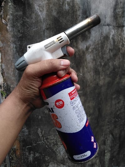An adult's hand is depicted holding a white "FLAME GUN" torch attached to a blue and white gas canister. The torch features a metallic nozzle, discolored and sooted at the tip from use, and a grey trigger. The gas canister, marked "NET 235gr" and displaying a red circular logo with East Asian characters, along with partial text including "PERKASA" and "GAS," shows surface dust or specks. The scene is set against a dark, weathered concrete or stucco wall with visible texture and minor cracks. The specific location within Mornington, Australia, cannot be determined from the provided image.