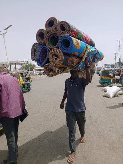 The image depicts an outdoor street scene in Kadoma, Zimbabwe, under bright daylight. A dark-skinned male, approximately in his 30s or 40s, is centrally positioned, walking towards the viewer's left. He is carrying a large stack of approximately 14-16 rolled-up, colorful floor mats or rugs on his head. The mats display various patterns, including floral designs. He is dressed in a dark blue short-sleeved polo shirt, dark grey cargo pants, and red-strapped flip-flops, with his left hand raised to steady the load. To the far left, another male in a purple long-sleeved shirt and dark pants walks away from the camera, holding a dark rectangular object. In the background, several auto-rickshaws (tuk-tuks) are visible, including a prominent green and yellow one on the left with a passenger in a yellow shirt, and another greenish one on the right next to white sacks resting on the ground. Further left, a street light pole with a solar panel and a structure resembling a covered station or market are present. Utility poles and wires are discernible on the right side of the background. The ground is paved, and the sky is light-colored, indicating a bright, possibly hazy, day.