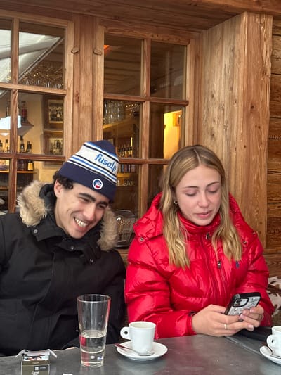 An interior scene captured in La Clusaz, France, depicts two individuals seated at a table in what appears to be a rustic establishment, possibly a restaurant or café. The background features light-colored wooden paneling and a large window with multiple panes, reflecting an interior bar area with bottles and glasses.

On the left, a male subject with dark, curly hair wears a black winter parka with a fur-lined hood and a blue, white, and light blue striped beanie featuring the text "fusalp" and a circular logo. He is smiling broadly, looking downwards and to his right, with his head slightly tilted.

To his right, a female subject with long, light brown hair wears a bright red puffy winter jacket, which appears to have a small embroidered logo on the chest. She is looking down intently at a white smartphone with a distinctive patterned case, holding it with both hands. She wears a ring on her left ring finger.

On the metallic grey table in front of them, a tall, clear glass contains water, positioned centrally. A white ceramic espresso cup with a saucer and spoon is directly in front of the female, and another similar cup is partially visible on the far right. A crumpled pack of Camel cigarettes rests on the far left side of the table.