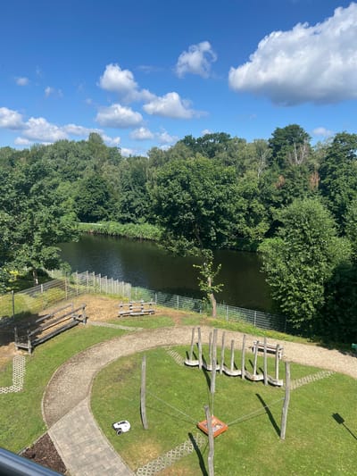 This is an aerial view of a park area next to a body of water, likely the Teltow Canal, in Teltow, Germany. A winding gravel path leads through a grassy area with several playground elements. To the left, there are two wooden benches and a rustic wooden structure. In the center, a row of wooden posts with curved seats attached suggests a balance beam or swing set. In the foreground, a white robotic lawnmower is parked on the grass. Beyond the park, the canal is visible, bordered by lush green trees on the opposite bank. The sky is blue with scattered white clouds.