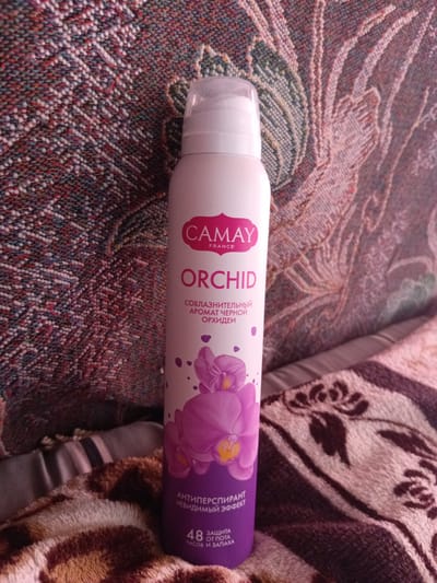 An unopened, off-white spray can of Camay Orchid deodorant, designed for women, stands upright on a multi-colored, textured blanket with a floral pattern. The can features the brand name "CAMAY FRANCE" in a pink heart-shaped logo at the top, followed by "ORCHID" in large, capitalized letters. Below the product name, in smaller text, are Russian words indicating "seductive aroma of black orchid." The lower half of the can displays a purple illustration of orchids and lists "ANTIPERSPIRANT INVISIBLE EFFECT" in Russian, along with "48 HOURS PROTECTION FROM SWEAT AND ODOR" also in Russian. The can is positioned in the center of the frame, against a blurred background of the blanket. The lighting suggests an indoor setting. There are no people or animals present in the image. The scene is static. The image was taken in Monastirische, Russia.