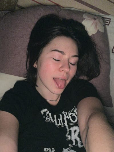 A young woman with dark hair lies on a pillow with closed eyes and tongue sticking out. She wears a black T-shirt with a white graphic design and a silver necklace. A tattoo is visible on her arm. The pillow has a floral pattern. The photo has a slightly grainy, vintage aesthetic.