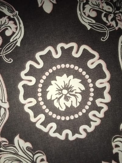 The image displays a tightly framed close-up of a dark grey textile fabric, exhibiting a repeating ornamental pattern in off-white and pale pink. A prominent central circular motif features a stylized off-white flower with multiple petals and a darker central core. This flower is immediately surrounded by a ring of uniformly spaced pale pink dots. Encircling the dotted ring is an undulating, ribbon-like border, rendered in off-white with pale pink edging. Partial elements of similar scrollwork and floral designs in off-white and pale pink are visible at the top-left, top-right, and bottom-left portions of the frame, consistent with a continuous pattern across the fabric surface. The textile material appears to be a fine weave with a slight sheen. No human subjects, actions, interactions, or specific environmental objects beyond the fabric itself are discernible within the frame. This observation was made in Umsobomvu Local Municipality, South Africa.