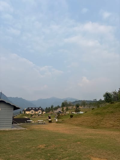 The image depicts a scenic outdoor setting with rolling hills, lush green grass, and a partly cloudy sky. In the foreground, a large grassy area dominates the lower half of the frame. A pathway leads from the lower left to a gray building with horizontal siding, partially visible on the far left.
Further into the scene, on a slightly elevated grassy slope, are several picnic tables and benches. A larger, light-colored building with a dark roof and stone accents is situated on a higher hill in the mid-ground. A few scattered trees, including pine trees, are present throughout the landscape. In the background, a range of hazy, tree-covered mountains forms the horizon. Several figures, appearing as people, are visible in the mid-ground, some standing near the picnic tables and others walking around. A light-blue vintage truck is parked on a road in the mid-right section of the image. The overall impression is a peaceful park-like environment. The location is identified as Babakan Madang, Indonesia.
