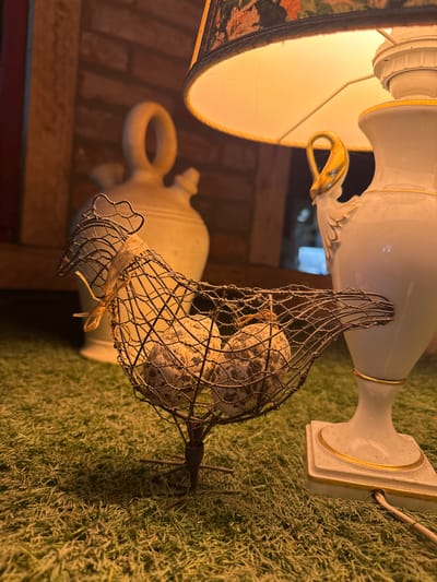 This media file features a close-up, vertically oriented shot of decorative objects arranged on a faux grass surface. The primary subjects are two cream-colored ceramic vases, one taller with gold trim and ornate detailing, and another shorter, jug-like vase with a loop handle.

Dominating the foreground is a wire cage containing what appear to be several speckled eggs. The cage itself is crudely formed, with a small strip of light-colored material tied around its opening. The eggs are clustered together within the cage.

Behind the wire cage, and partially obscured, is a lamp with a patterned lampshade, emitting a warm, yellow light that illuminates the scene. In the background, the base of the taller vase is visible, resting on the textured green surface. The overall impression is of a still life arrangement, possibly a decorative display for a garden or indoor setting.

The lighting suggests it might be evening or indoors, with artificial light providing illumination. There are no people visible, and the focus is entirely on the inanimate objects. The textures of the ceramic, wire, eggs, and faux grass are all discernible. There is no visible text in the image.