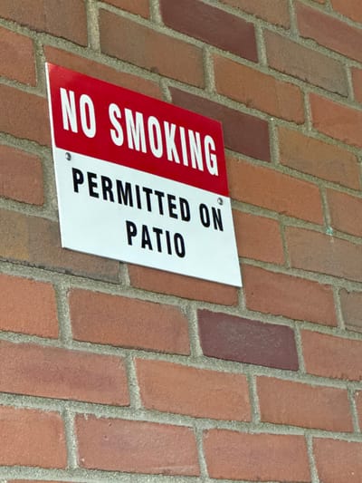 A white rectangular sign with red and black text is affixed to a reddish-brown brick wall. The sign features a red upper band with the white capitalized text "NO SMOKING". Below this, on the white background, is the black capitalized text "PERMITTED ON PATIO". The sign is secured to the wall by four visible silver fasteners, one in each corner, though the bottom right fastener is partially obscured by the image crop. The brick wall exhibits varying shades of red and brown bricks with light gray mortar lines. The scene is located in the City of Binghamton, United States. No individuals or specific actions are depicted.