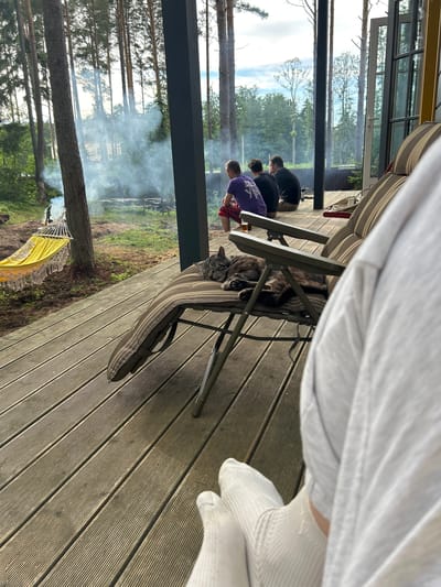 A group of three men are seated on a wooden deck outside a building, with a cat sleeping on a lounge chair. One man wears a purple t-shirt and red and black plaid shorts, another a black t-shirt, and the third a dark t-shirt. Smoke is visible in the background, suggesting a bonfire or grill. A yellow hammock is strung between trees to the left of the deck. The scene appears to be a rural or semi-rural setting, possibly near Shepelyovo, Russia, given the tall pine trees and natural landscape. The perspective is from someone on the deck, looking towards the group and the outdoor environment.
