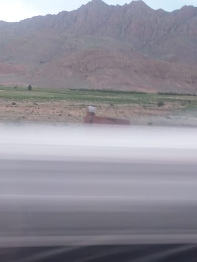 The image captures a vast, mountainous landscape, likely viewed from a moving vehicle. The scene is dominated by a rugged, reddish-brown mountain range with sparse green vegetation, extending across the left and upper parts of the frame. In the middle ground, a narrow strip of land, varying in shades of green and brown, suggests either a road, a track, or cultivated fields running parallel to the mountains. A small, light-colored structure, possibly a building or shelter, is situated on this strip of land towards the center. The right side of the image is heavily blurred with vertical streaks of white and grey, indicative of motion blur from a window, or an obstruction such as a curtain or vehicle pillar. The lighting suggests it is daytime with clear weather conditions. The overall setting is an open, natural environment consistent with a mountainous region. No people or visible text are discernible in the image.