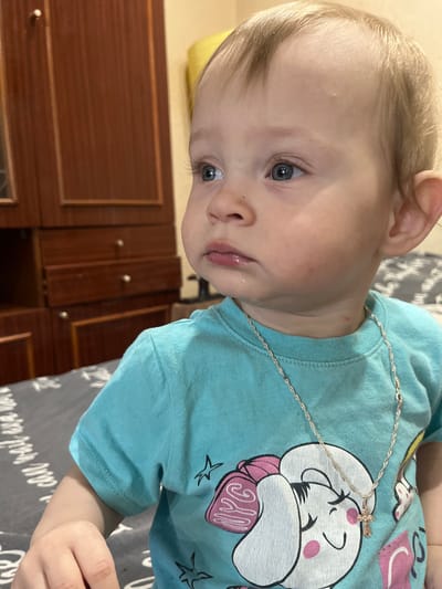 A close-up, eye-level shot shows a Caucasian toddler with blonde hair and blue eyes looking to the left, with lips parted and a slight drool on the chin. The toddler wears a light blue t-shirt with a graphic of a cartoon character. A thin silver necklace with a small cross pendant rests on the t-shirt. In the background, a wooden wardrobe with three drawers is visible, with a slightly blurred bed with a gray patterned sheet. The location is identified as Alatyr, Russia.