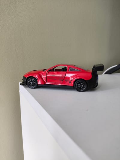 A red toy sports car with a black spoiler is positioned on a white surface. The car is seen from a side profile, with its left side facing the viewer. The surface it rests on is angled away from the camera. In the background, a textured wall is visible, with a gradient of light and shadow. The scene is indoors and appears to be a still life. There is no indication of the city of Kaluga, Russia, or any people present.